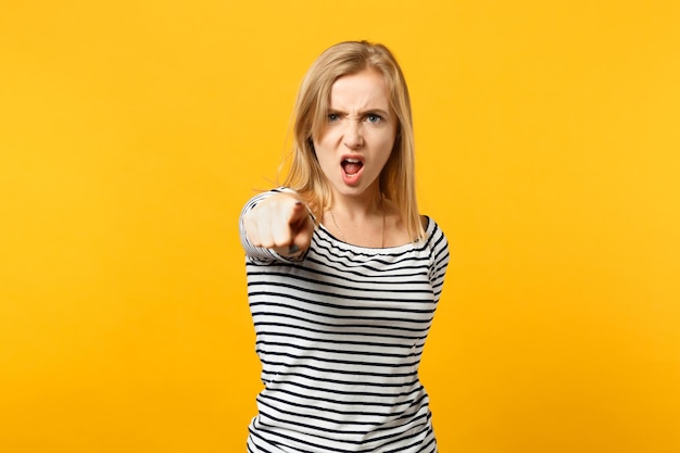 Angry young woman in striped clothes pointing index finger on camera, screaming swearing isolated on yellow orange background in studio. People sincere emotions lifestyle concept. Mock up copy space.