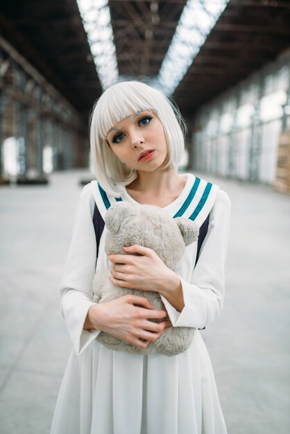 Блондинка в стиле аниме обнимает плюшевого мишку. Женщина-косплей, японская культура, кукла с игрушкой на заброшенной фабрике