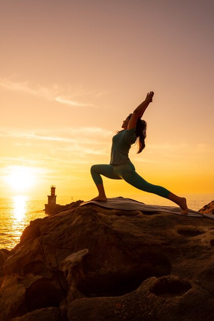 Anjaneyasana een vrouw die meditatie en yoga-oefeningen doet op een rots bij zonsondergang naast een vuurtoren in de zee, gezond en naturistisch leven
