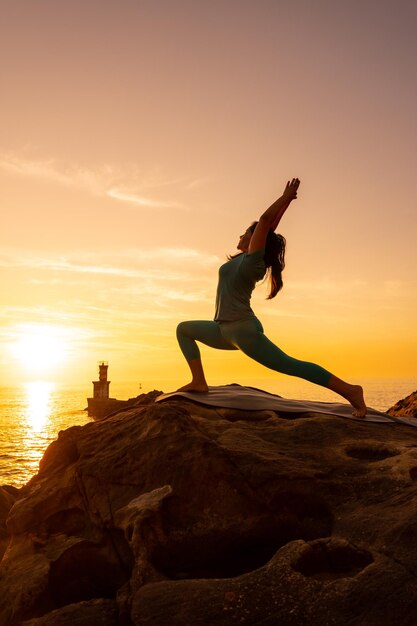 Anjaneyasana Een vrouw die meditatie en yoga-oefeningen doet op een rots bij zonsondergang naast een vuurtoren in de zee, gezond en naturistisch leven