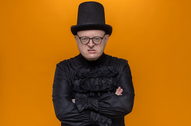 Annoyed adult man with top hat and glasses in black gothic shirt crossing arms and looking 