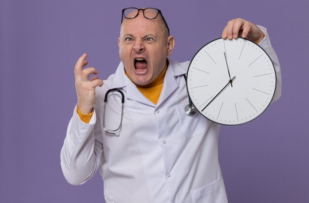 Annoyed adult slavic man with optical glasses in doctor uniform with stethoscope holding clock and yelling at someone looking at side