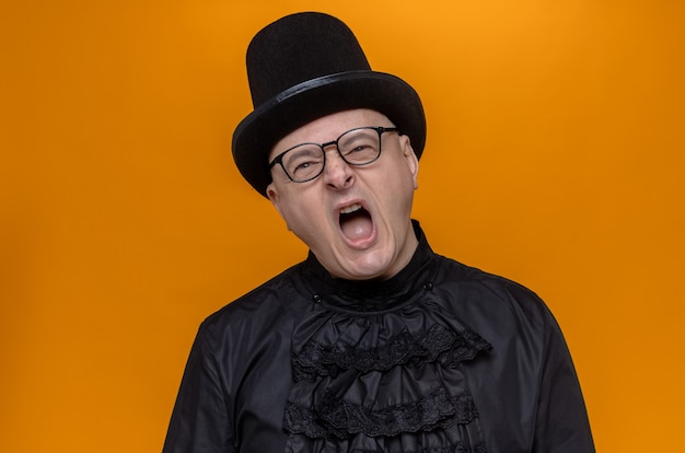 Annoyed adult slavic man with top hat and optical glasses in black gothic shirt yelling at someone looking at front