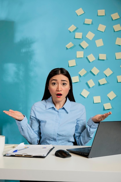 Annoyed asian woman sitting at office desk making hand gesture asking for explanation. Female employee looking at statistics results at workplace unable to believe what is happening.