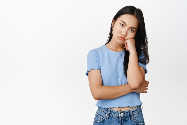 Annoyed or bored asian girl lean on hand looking pissed off and offended at camera standing in blue tshirt over white background