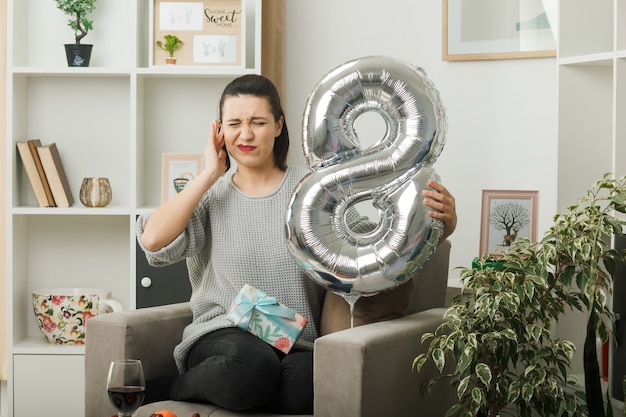 Annoyed closed ear beautiful girl on happy women day holding number eight balloon sitting on armchair in living room