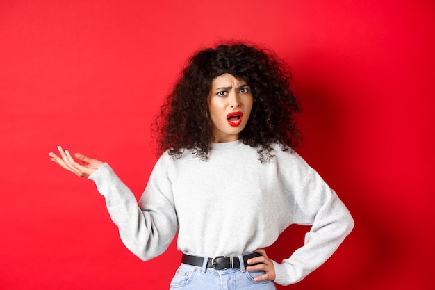 Donna caucasica infastidita e confusa con i capelli ricci, alzando la mano e litigando, litigando, non riesco a capire qualcosa, in piedi su sfondo rosso.