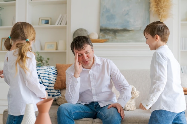 An annoyed father sitting on sofa at home in living room distracted from noise and touching his temp...