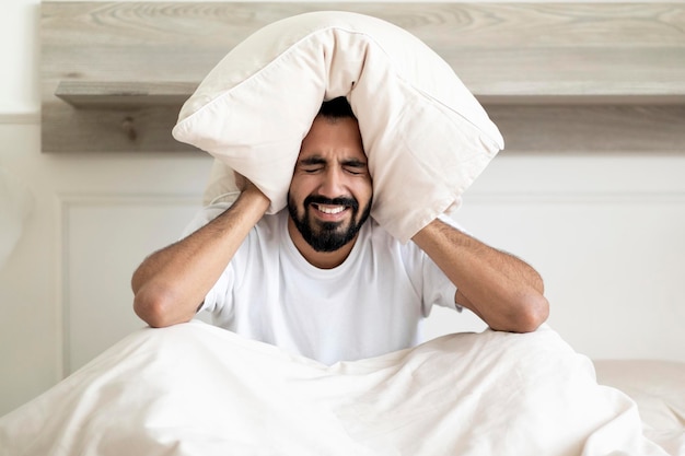 Annoyed indian guy covering ears with pillow sitting on bed at home