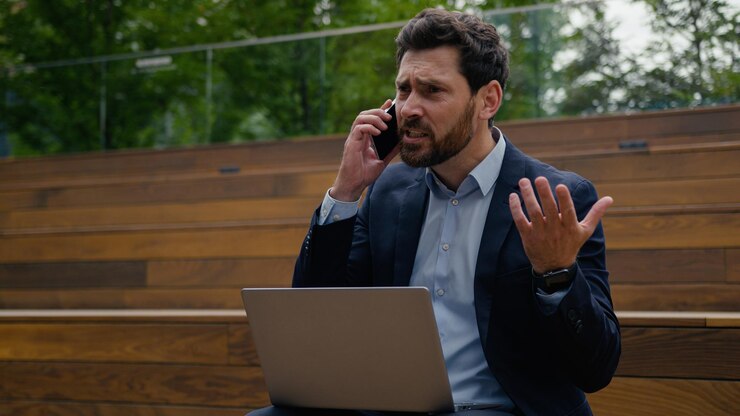 Photo annoyed mad angry man caucasian businessman furious business leader work remote laptop city outdoors male guy talk mobile phone quarrel shout fight scream conflict finish call throwing cellphone away