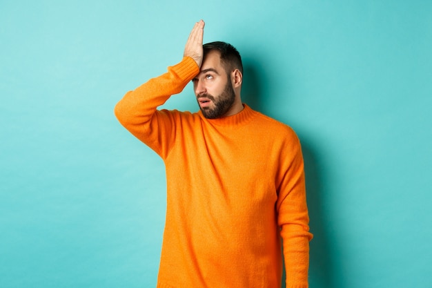 Uomo infastidito alzare gli occhi al cielo e fare facepalm, in piedi su sfondo turchese.