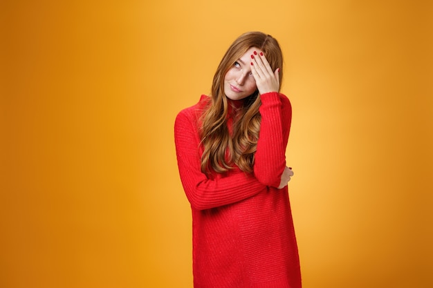 Annoyed and pissed embarrassed woman hiding behind hand making facepalm gesture looking away with irritated humiliated gesture, standing fed up and intense against orange background.