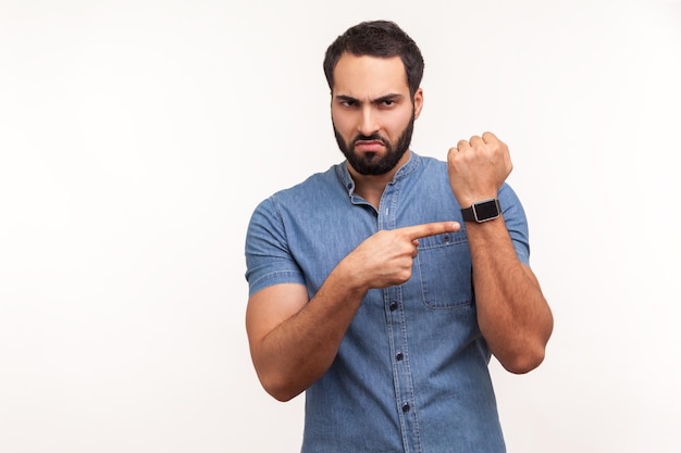 Annoyed punctual man with beard in blue shirt pointing finger at wrist watch mock up display and expressing dissatisfaction with late time deadline Indoor studio shot isolated on white background