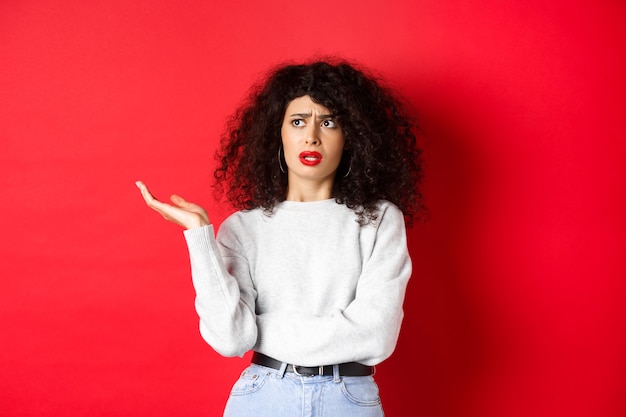 Annoyed and puzzled young woman with curly hair, raising hands up and looking aside, cant understand something strange, standing on red wall