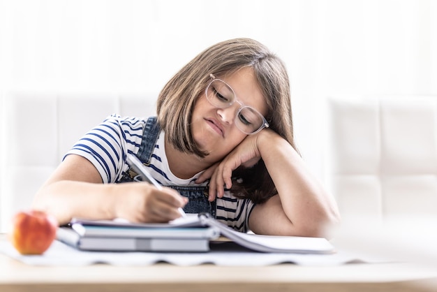 Annoyed schoolgirl with eyewear writes homework leaning against her hand