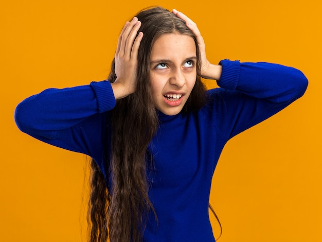 Annoyed teenage girl keeping hands on head looking up isolated on orange wall