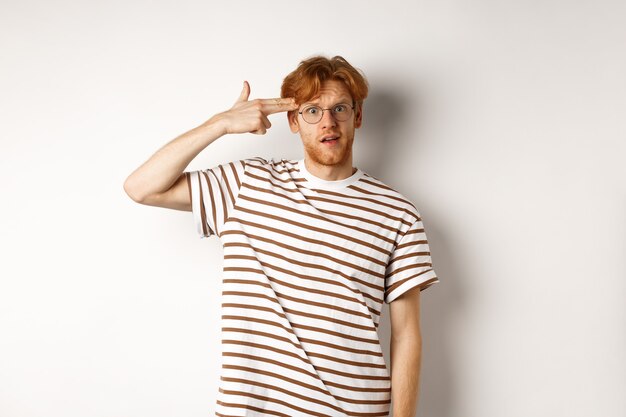 Annoyed teenage redhead guy making finger gun near head, staring bothered and irritated at camera, white background.