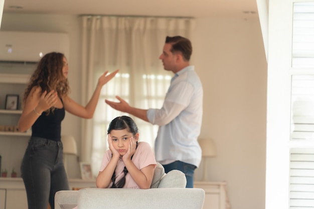 Annoyed and unhappy young girl look at camera trapped in middle of tension by her parent argument in living room Unhealthy domestic lifestyle and traumatic childhood develop to depressionSynchronos