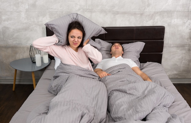 Annoyed wife blocking her ears by a pillow from noise of husband snoring while sleeping in the bed in the bedroom in loft style with grey colors