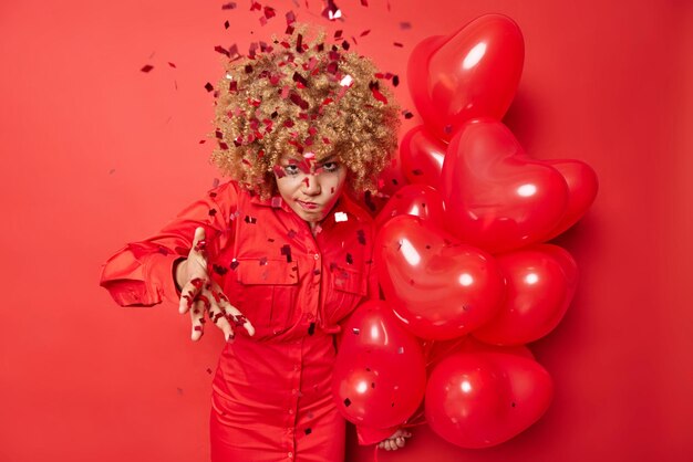 Annoyed woman doesnt like surprise received on birthday wears\
dress holds bunch of heart balloons angry with falling confetti on\
her has irritated face expression isolated over red background