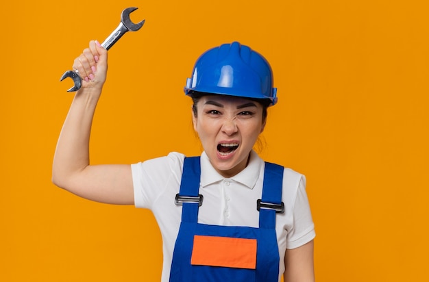 Annoyed young asian builder girl with blue safety helmet stands with raised hand holding workshop key