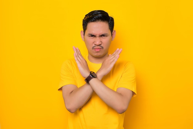 Annoyed young asian man in casual tshirt crossing hands making\
stop gesture and demonstrates rejection isolated on yellow\
background people lifestyle concept