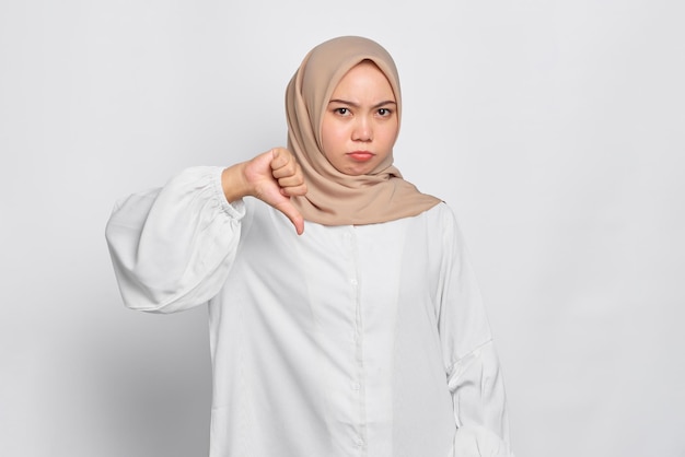 Annoyed young Asian Muslim woman showing thumbs down isolated over white background