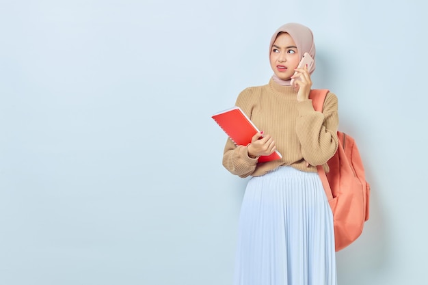 携帯電話で話し、学校のコンセプトに戻って白い背景で隔離の本を保持しているバックパックと茶色のセーターでイライラする若いアジアのイスラム教徒の女性の学生