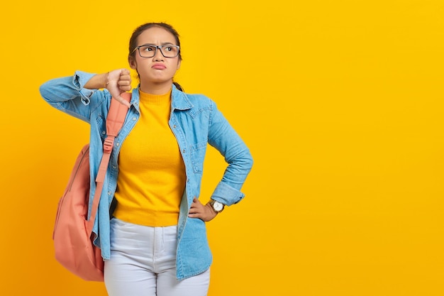 Annoyed young asian student in denim outfit with backpack\
showing thumbs down with finger isolated on yellow background\
education in high school university college concept