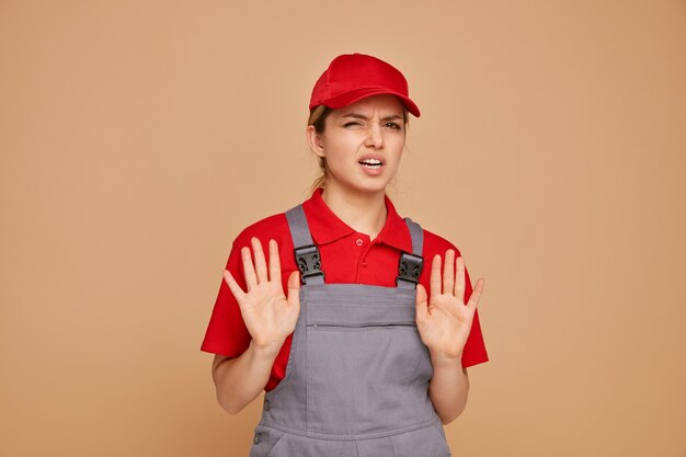 Annoyed young female construction worker wearing uniform and cap doing refusal gesture 