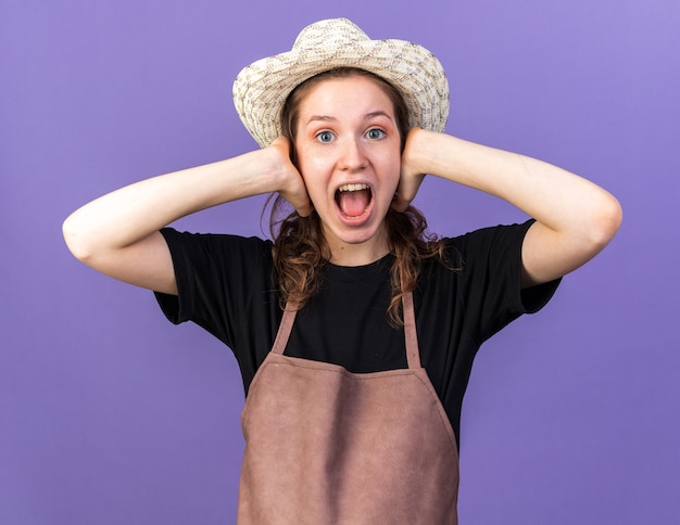 Annoyed young female gardener wearing gardening hat covered ears with hands isolated on blue wall