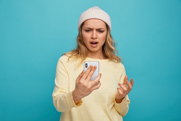 Foto ragazza infastidita che indossa cappello invernale tenendo e guardando il telefono cellulare tenendo la mano in aria