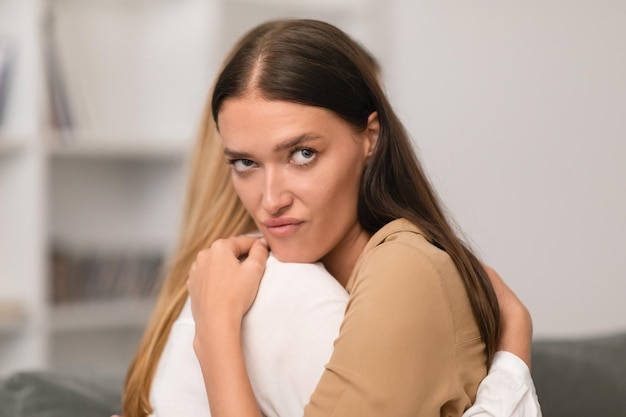 annoyed young lady embracing her girlfriend with displeased expression indoor
