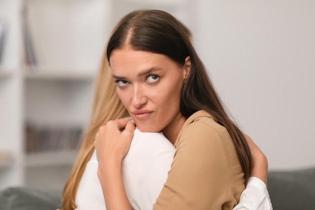 Annoyed young lady embracing her girlfriend with displeased expression indoor