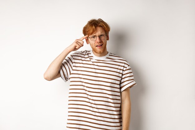 Annoyed young man with red hair scolding person for being stupid, pointing at head and staring at camera, white background.