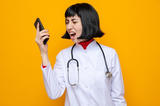 Annoyed young pretty caucasian woman in doctor uniform with stethoscope holding and looking at phone 