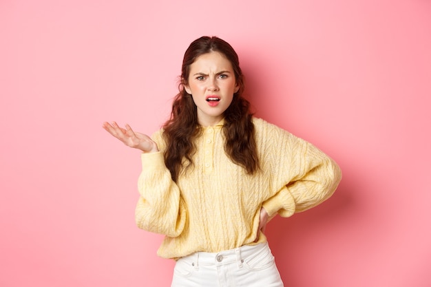 Annoyed young woman complaining at something strange, staring confused, raising one hand with puzzled face, standing over pink wall