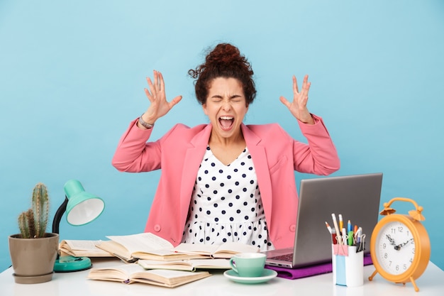 Photo annoyed young woman screaming and throwing up hands while working at desk isolated over blue wall
