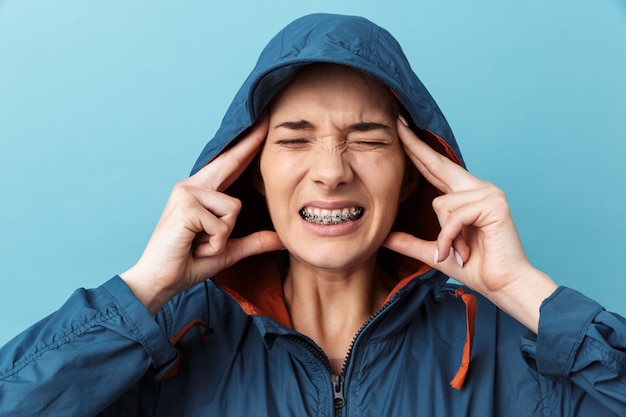 Annoyed young woman wearing a jacket standing isolated over blue wall, suffering from a headache