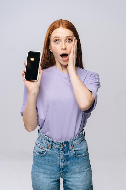 Annoyed young woman wearing stylish casual clothes holding cell phone with black empty mobile screen Pretty lady model with red hair emotionally showing facial expressions in studio copyspace