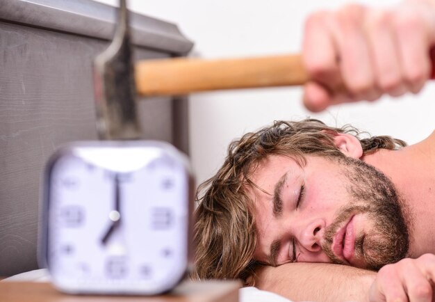 Annoying ringing alarm clock. Man bearded annoyed sleepy face lay pillow near alarm clock. Guy knocking with hammer alarm clock ringing. Break discipline regime. Annoying sound. Stop ringing.