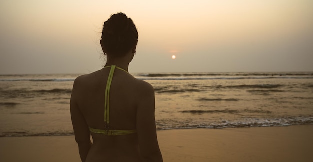 Anonieme vrouw in de buurt van zee tijdens zonsondergang Achteraanzicht van onherkenbare vrouw in badmode staande op strand in de buurt van wuivende zee in de avond op resort