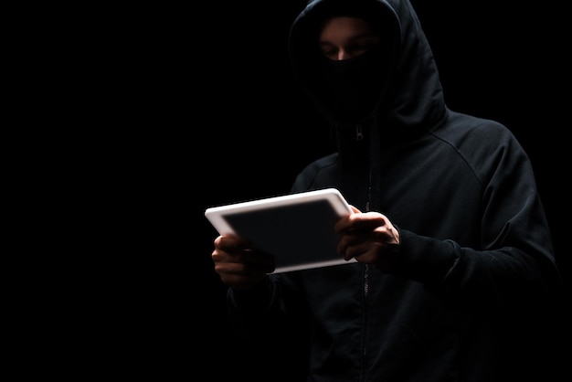 Anonymous hacker in hood using digital tablet isolated on black