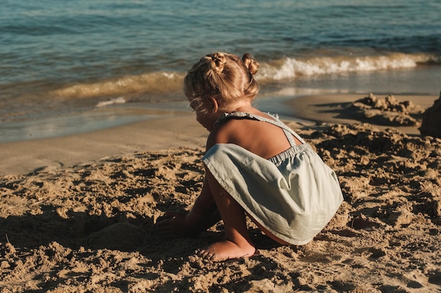 Анонимная маленькая девочка в повседневной одежде играет с песком у моря на пляже в солнечный день