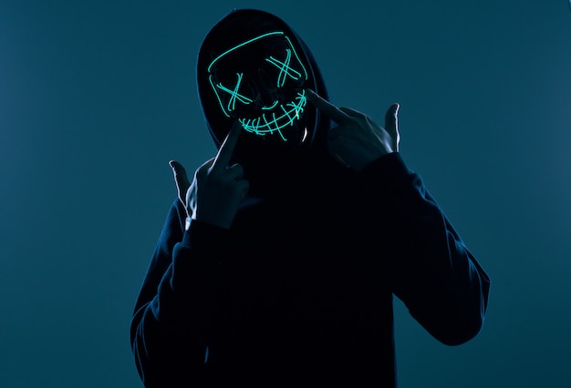 Anonymous man in black hoodie hiding his face behind a neon mask