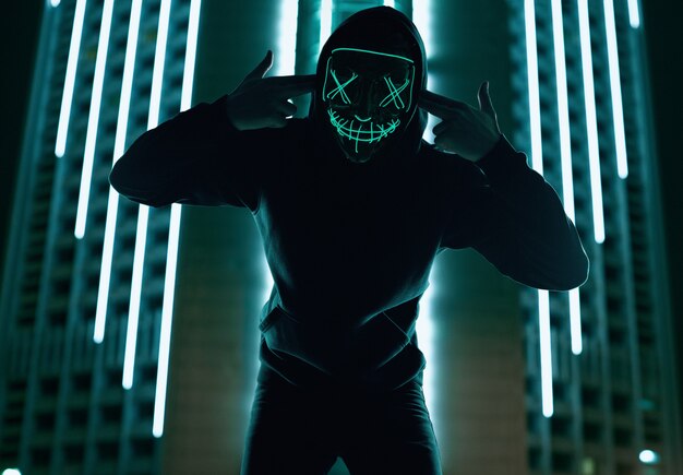 Anonymous man in black hoodie hiding his face behind a neon mask