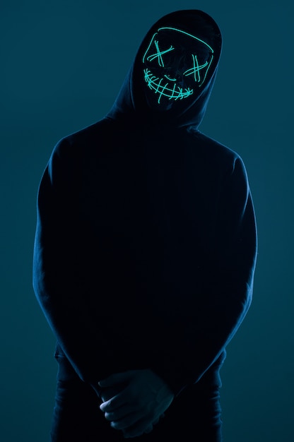 Anonymous man in black hoodie hiding his face behind a neon mask