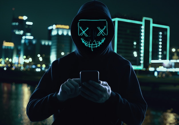 Анонимный мужчина в черной балахоне и неоновой маске взламывает смартфон