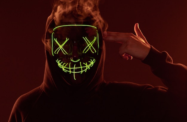 Photo anonymous man hiding his face behind neon mask in a colored smoke