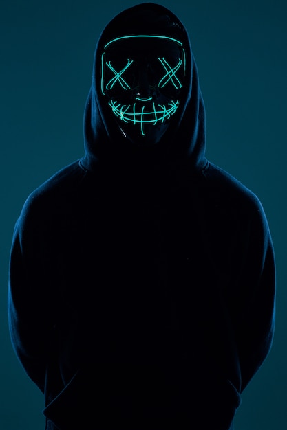 Анонимный мужчина в черной толстовке с капюшоном прячет лицо за неоновой маской
